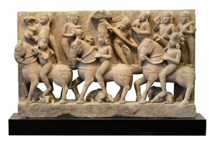 This is a royal procession scene from a medieval, Indian temple.  The central rider wears an embellished helmet, or crown, that is indicative of his royal status.  He is accompanied on either side by two companions, both of whom turn to face him out of deference.  On the frieze behind the riders, a throng of supporters watches the procession.  The carver has conveyed a sense of depth both by placing the followers on a ledge behind the main figures and by making these secondary figures smaller in scale.  While this technique lacks the sophistication of, for instance, classical Greco-Roman friezes, the artist still achieves a sense of depth and perspective while providing space in which to depict the horses and central figures in a voluminous manner.  In addition, the carver reveals particular skill in the detail work on the horses’ saddles and harnesses and on the riders’ boots and armored leggings.